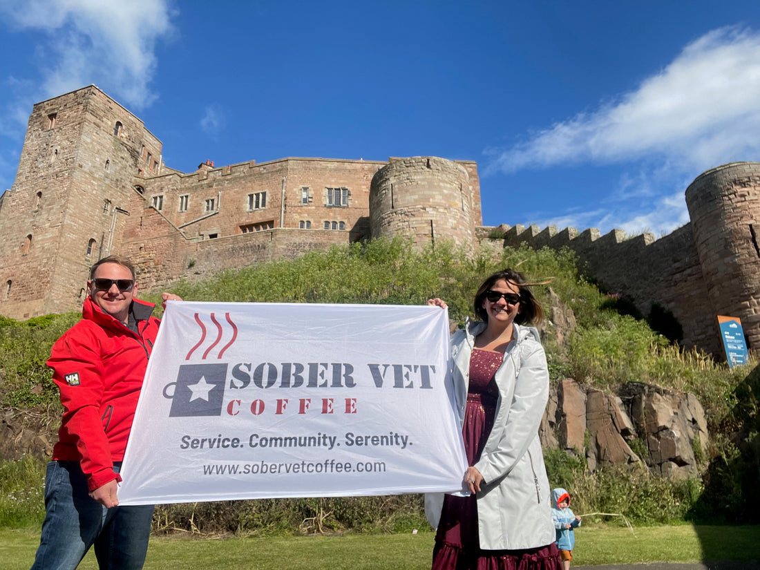 David and Kristina (co-founders) stand in front of Bamburgh Castle holding a Sober Vet Coffee banner